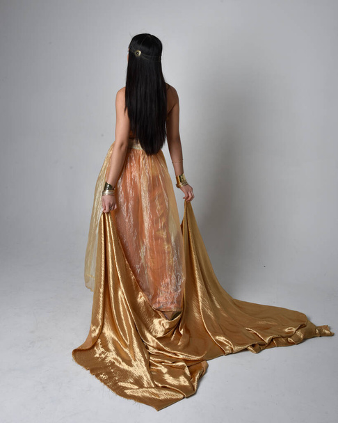 Full length portrait of pretty young asian woman wearing golden Arabian robes like a genie, standing pose holding flowing fabric, isolated on studio background. - Fotó, kép