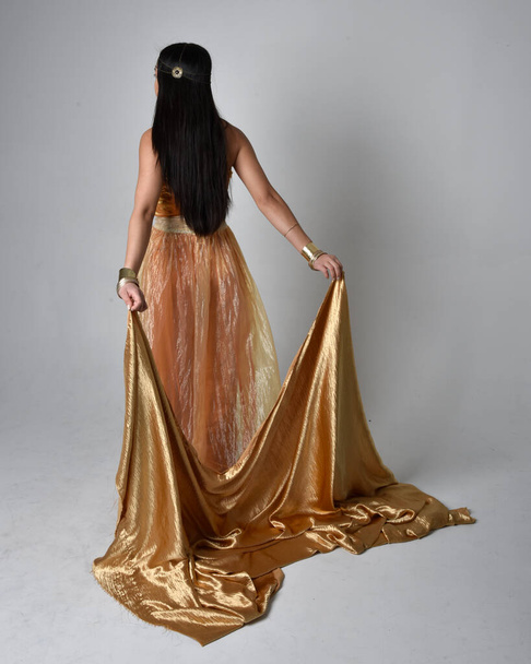 Full length portrait of pretty young asian woman wearing golden Arabian robes like a genie, standing pose holding flowing fabric, isolated on studio background. - Photo, Image