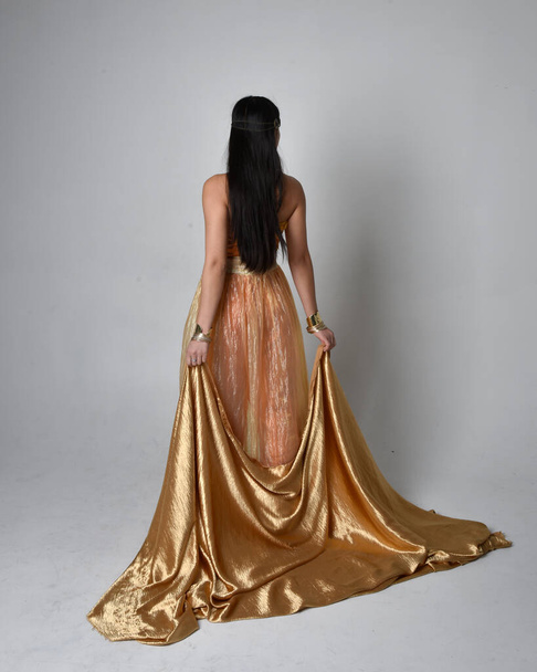 Full length portrait of pretty young asian woman wearing golden Arabian robes like a genie, standing pose holding flowing fabric, isolated on studio background. - Φωτογραφία, εικόνα