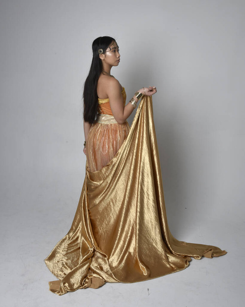 Full length portrait of pretty young asian woman wearing golden Arabian robes like a genie, standing pose holding flowing fabric, isolated on studio background. - Foto, Imagem