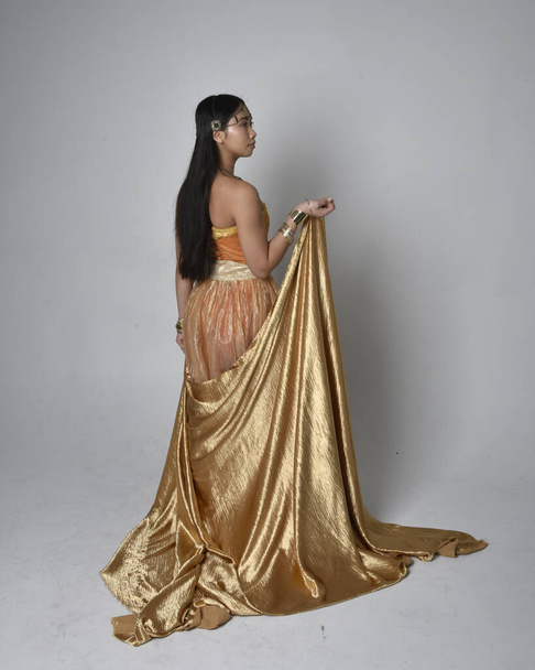 Полный портрет красивой молодой азиатской женщины в золотых арабских одеждах, как джинн, стоящей в позе держа текущую ткань, изолированные на студийном фоне. - Фото, изображение