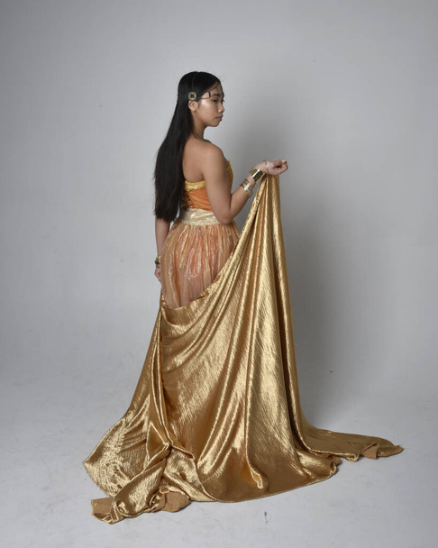 Full length portrait of pretty young asian woman wearing golden Arabian robes like a genie, standing pose holding flowing fabric, isolated on studio background. - Foto, Imagen
