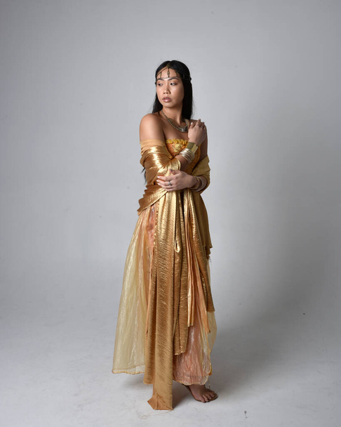 Altın Arap cübbesi giymiş güzel Asyalı bir kadının tam boy portresi. Stüdyo arka planında izole edilmiş, akan kumaşı tutan bir cin gibi duruyor.. - Fotoğraf, Görsel