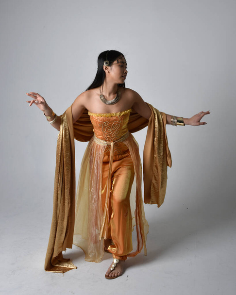 Full length portrait of pretty young asian woman wearing golden Arabian robes like a genie, standing pose holding flowing fabric, isolated on studio background. - Фото, зображення