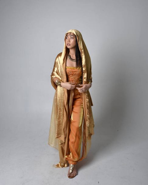 Portret pięknej młodej azjatyckiej kobiety w złotych arabskich szatach, jak dżin, stojącej w pozycji stojącej z płynącą tkaniną, odizolowanej na tle studia. - Zdjęcie, obraz