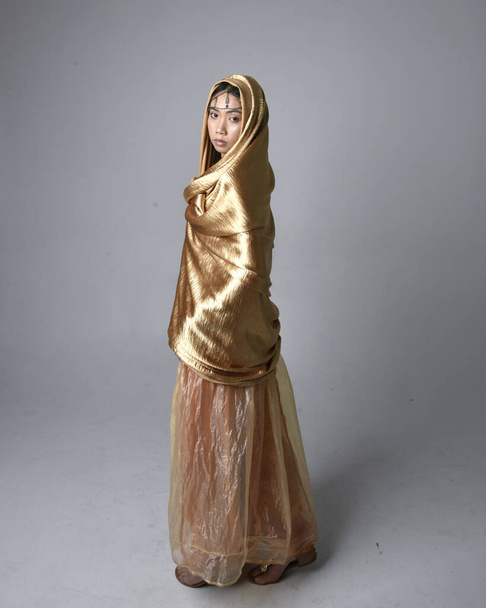 ジーニーのような黄金のアラビア語のローブを身に着けているかなり若いアジアの女性の完全な長さの肖像画,流れるような布を保持立ってポーズ,スタジオの背景に孤立. - 写真・画像