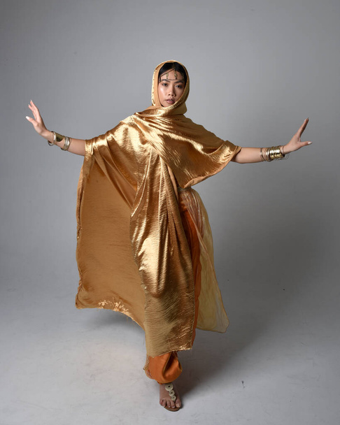 Full length portrait of pretty young asian woman wearing golden Arabian robes like a genie, standing pose holding flowing fabric, isolated on studio background. - Foto, Bild