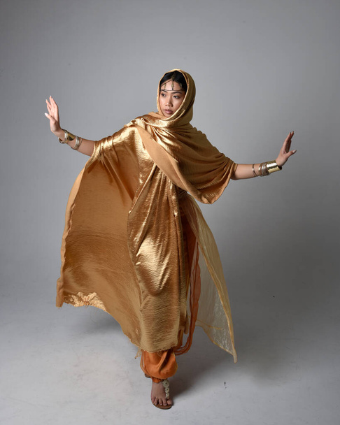 Full length portrait of pretty young asian woman wearing golden Arabian robes like a genie, standing pose holding flowing fabric, isolated on studio background. - Zdjęcie, obraz