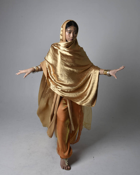 Full length portrait of pretty young asian woman wearing golden Arabian robes like a genie, standing pose holding flowing fabric, isolated on studio background. - Foto, immagini