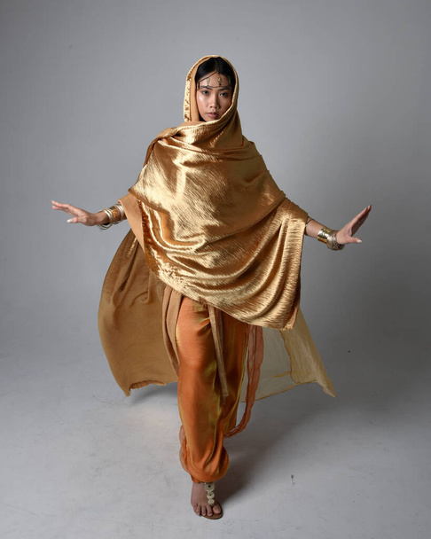 Full length portrait of pretty young asian woman wearing golden Arabian robes like a genie, standing pose holding flowing fabric, isolated on studio background. - Valokuva, kuva