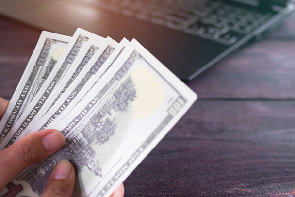Finger holding bank note 100 dollars with keyboard laptop computer blur on wooden table. planing earn money investment and saving money future for 2022 new year. finance and business. count money pay - Photo, Image