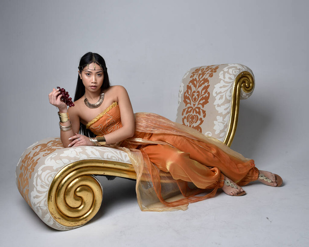 Full length portrait of pretty young asian woman wearing golden Arabian robes like a genie, seated pose on lounge, isolated on studio background. - Photo, Image
