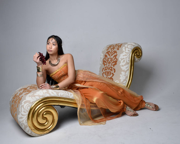 Full length portrait of pretty young asian woman wearing golden Arabian robes like a genie, seated pose on lounge, isolated on studio background. - 写真・画像