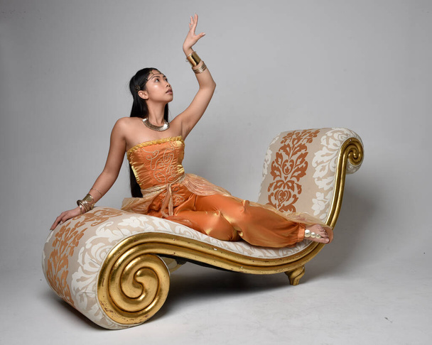 Ganztägiges Porträt einer hübschen jungen asiatischen Frau, die goldene arabische Roben wie ein Genie trägt, auf Lounge sitzend, isoliert auf Studiohintergrund. - Foto, Bild