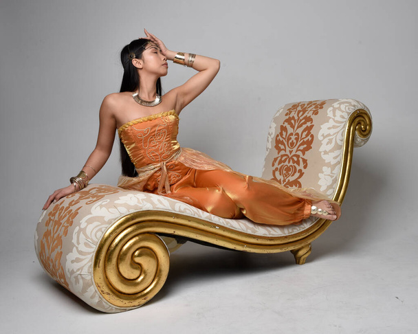 Ganztägiges Porträt einer hübschen jungen asiatischen Frau, die goldene arabische Roben wie ein Genie trägt, auf Lounge sitzend, isoliert auf Studiohintergrund. - Foto, Bild