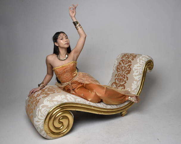 Täyspitkä muotokuva kaunis nuori aasialainen nainen yllään kultainen Arabian kylpytakit kuin henki, istuu poseeraa lounge, eristetty studio tausta. - Valokuva, kuva