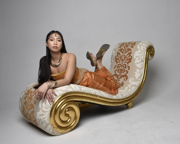 Teljes hossz portré csinos fiatal ázsiai nő visel arany arab köntös, mint egy dzsinn, ülő póz társalgóban, elszigetelt stúdió háttér. - Fotó, kép