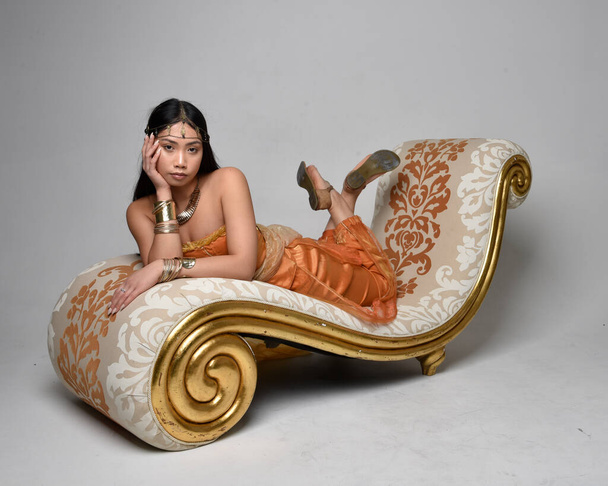 Täyspitkä muotokuva kaunis nuori aasialainen nainen yllään kultainen Arabian kylpytakit kuin henki, istuu poseeraa lounge, eristetty studio tausta. - Valokuva, kuva