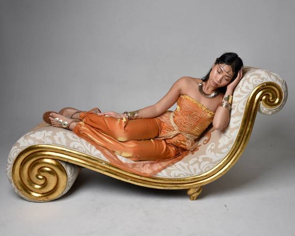 Full length portrait of pretty young asian woman wearing golden Arabian robes like a genie, seated pose on lounge, isolated on studio background. - 写真・画像