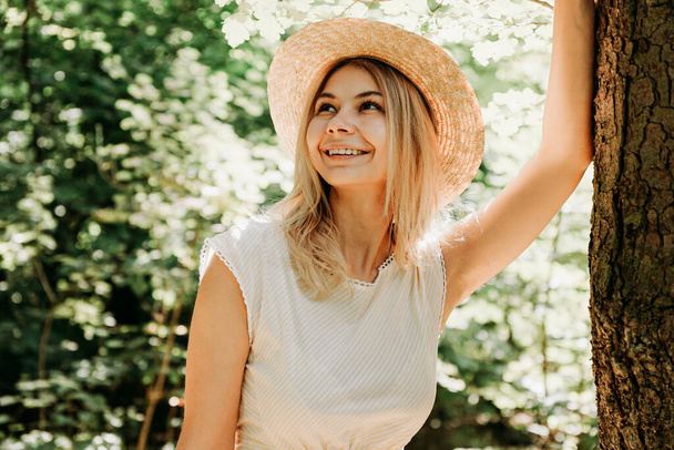 Beautiful girl in a straw hat and stylish clothes stands in a park near a tree - Fotoğraf, Görsel