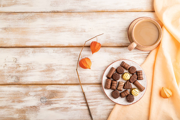 Schokoladenbonbons mit Tasse Kaffee und Physalis-Blüten auf weißem Holzgrund und orangefarbenem Textil. Draufsicht, flache Lage, Kopierraum. - Foto, Bild