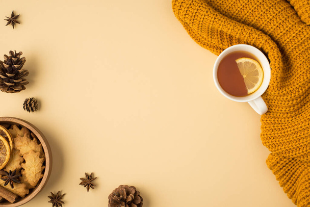 Foto de vista superior de suéter de punto amarillo taza de té con conos de pino rodaja de limón y tazón con galletas en forma de hoja y rodajas de limón secas sobre fondo naranja pastel aislado con espacio vacío - Foto, imagen
