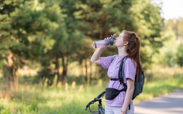 Redhead girl rides a bicycle in a beautiful summer forest. Woman cyclist drinks water from a reusable bottle - Fotografie, Obrázek