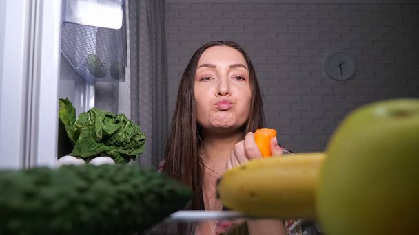 Woman vegan looks for snacks and takes carrot from fridge - Фото, изображение