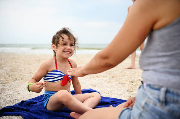 Αξιολάτρευτο κοριτσάκι ξεκουράζεται στην παραλία, παίζει παιχνίδια με τη μητέρα της, χαριτωμένο χαμογελώντας ποζάροντας στην κάμερα με φόντο την αμμώδη παραλία. Έννοιες για χαρούμενες και ξέγνοιαστες καλοκαιρινές διακοπές στην παραλία - Φωτογραφία, εικόνα