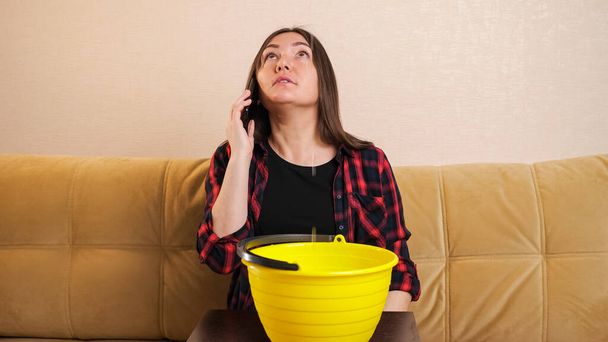Lady calls plumber collecting water into bucket on sofa - Foto, Imagen