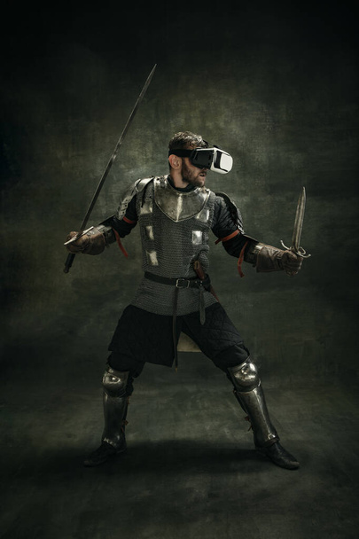 Portrait of one brutal bearded man, medeival warrior or knight in VR glasses with sword over dark background. - Фото, изображение