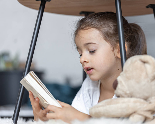 Portrait d'une mignonne petite fille lisant un livre à la maison, allongée sur le sol avec son jouet préféré. - Photo, image