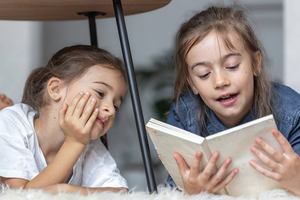 Дві маленькі сестри весело читають книгу разом, лежачи на підлозі у своїй кімнаті
. - Фото, зображення