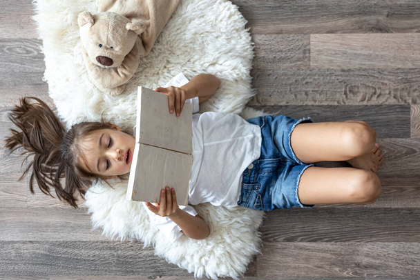 Το παιδί διαβάζει ένα βιβλίο ξαπλωμένο σε ένα άνετο χαλί στο σπίτι με το αγαπημένο του αρκουδάκι.. - Φωτογραφία, εικόνα