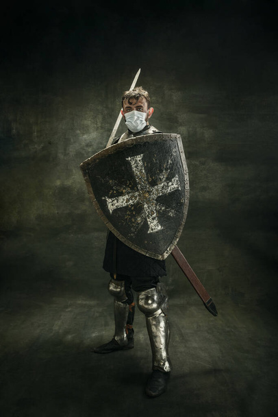 Ένας κτηνώδης γενειοφόρος άνδρας, μεταδιδόμενος πολεμιστής ή ιππότης με ιατρική μάσκα με ασπίδα σε σκούρο φόντο.. - Φωτογραφία, εικόνα