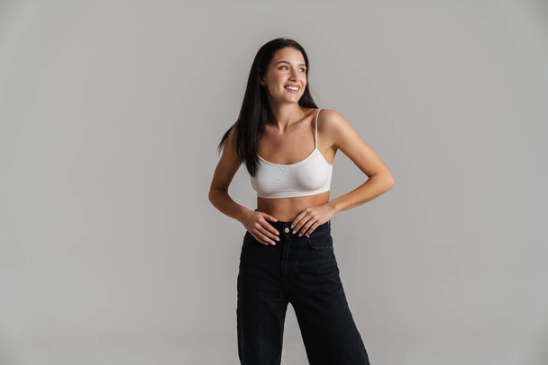 Smiling young white woman with long brunette hair wearing crop top standing posing over gray background - Fotografie, Obrázek