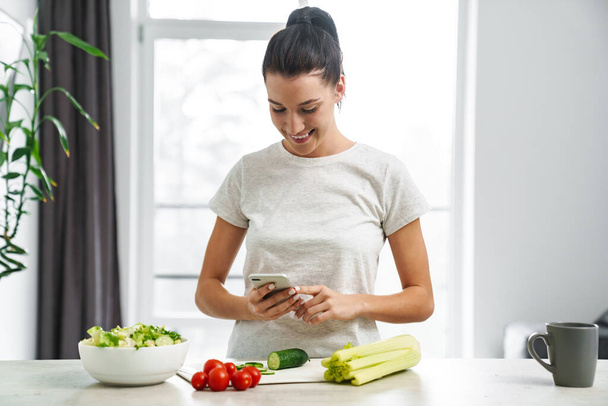 European smiling woman using mobile phone and making salad in kitchen at home - Zdjęcie, obraz