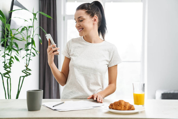 Европейская улыбающаяся женщина, пользующаяся мобильным телефоном во время завтрака на кухне дома - Фото, изображение