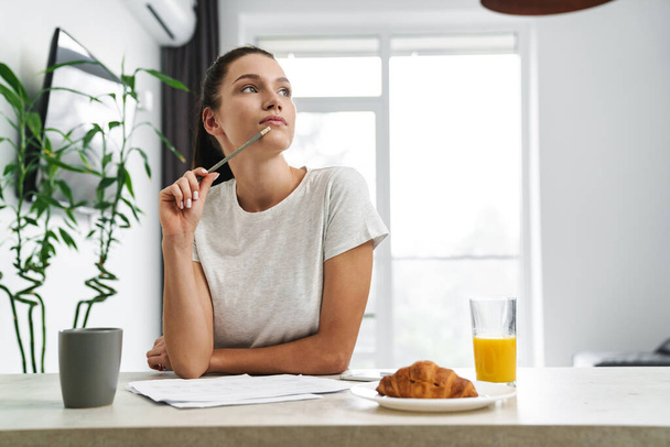 European thinking woman writing down notes while having breakfast in kitchen at home - Photo, image