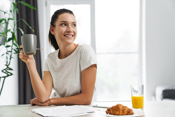 Ευρωπαία νεαρή γυναίκα πίνει καφέ ενώ τρώει πρωινό στην κουζίνα στο σπίτι - Φωτογραφία, εικόνα