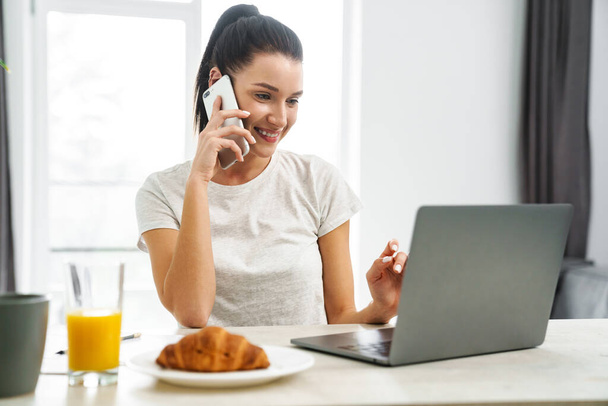 Smiling woman talking on cellphone and using laptop while having breakfast in kitchen at home - Φωτογραφία, εικόνα