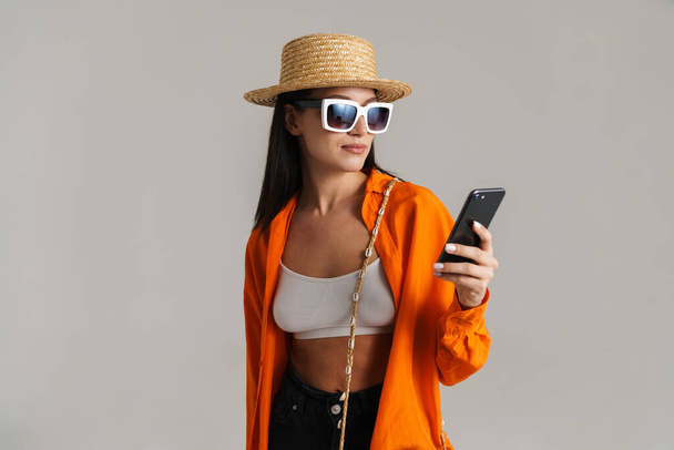 Young brunette woman in hat and sunglasses using mobile phone isolated over grey background - Fotografie, Obrázek