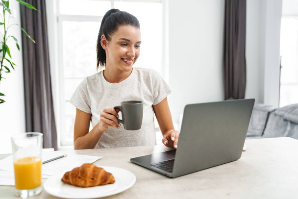 European smiling woman using laptop while drinking coffee in kitchen at home - Foto, Imagen