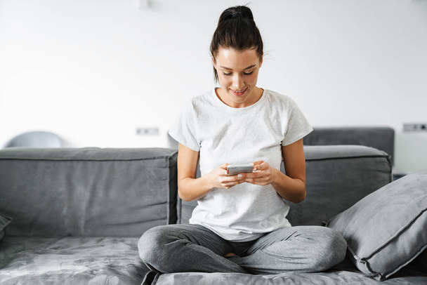 European smiling woman using mobile phone while sitting on couch at home - Foto, imagen