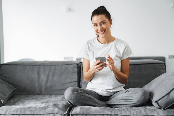 European smiling woman using mobile phone while sitting on couch at home - Photo, Image