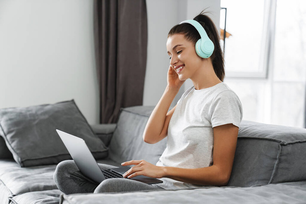 European smiling woman using headphones and laptop while sitting on couch at home - Fotoğraf, Görsel