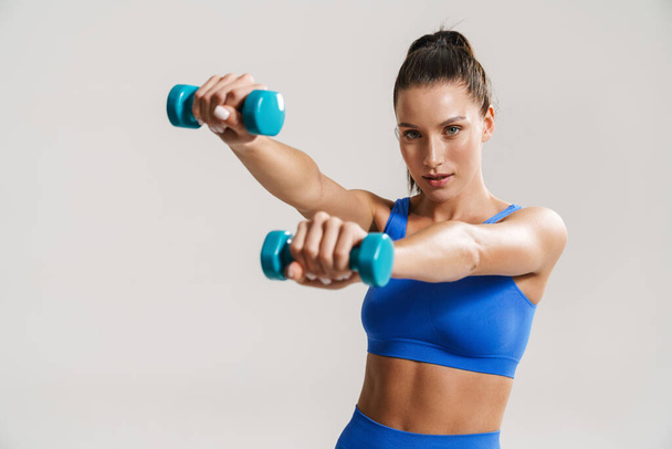 Young white fitness woman in sportswear with ponytail standing over white wall background, lifting small weights - Foto, afbeelding