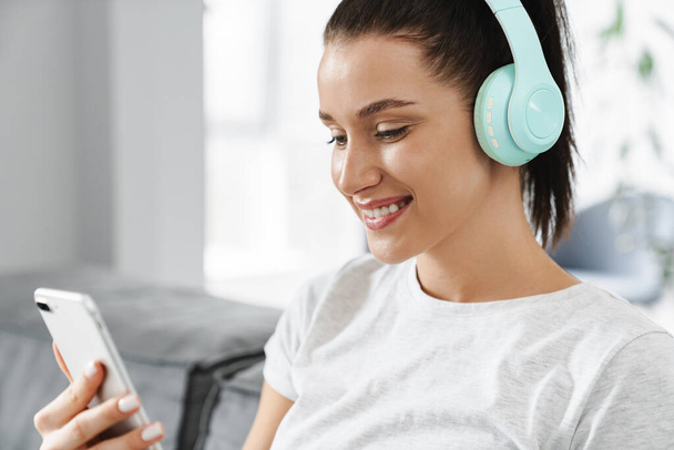 Smiling woman using cellphone and headphones while sitting on couch at home - Foto, immagini