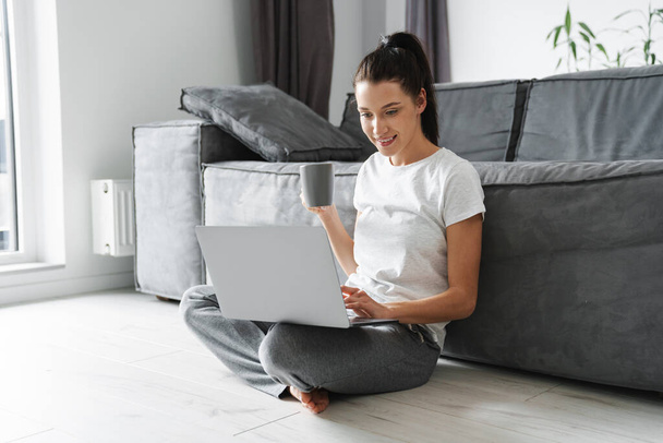 Smiling woman drinking coffee and using laptop while sitting on floor at home - Foto, immagini