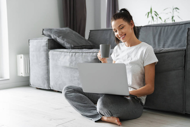 笑顔の女性はコーヒーを飲みながら、自宅で床に座っている間にラップトップを使用して - 写真・画像